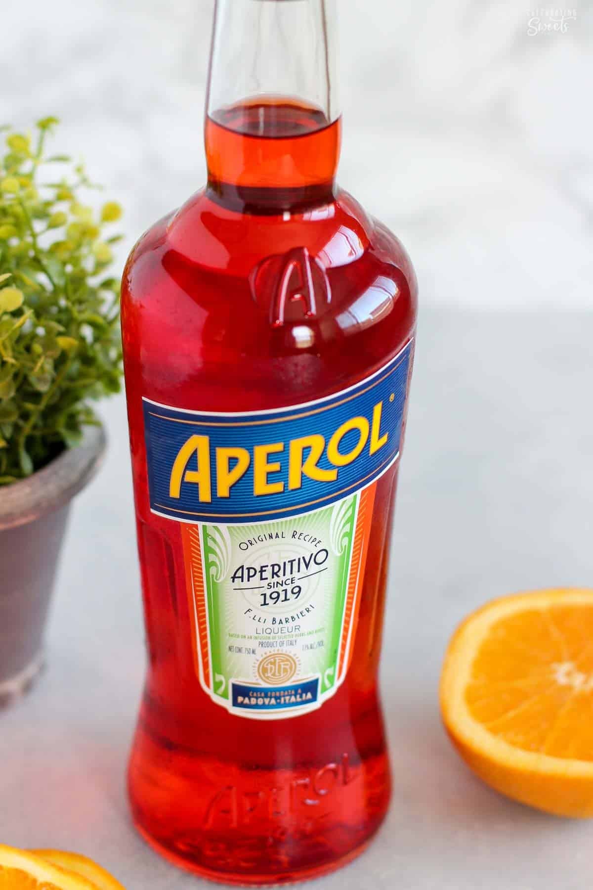 Bottle of aperol next to an orange and a small potted plant.