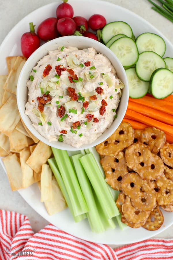 Bowl of dip topped with artichokes and sun dried tomatoes served with vegetables and pretzels.