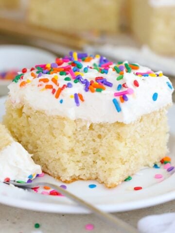 Slice of vanilla cake with vanilla frosting and sprinkles on a white plate.