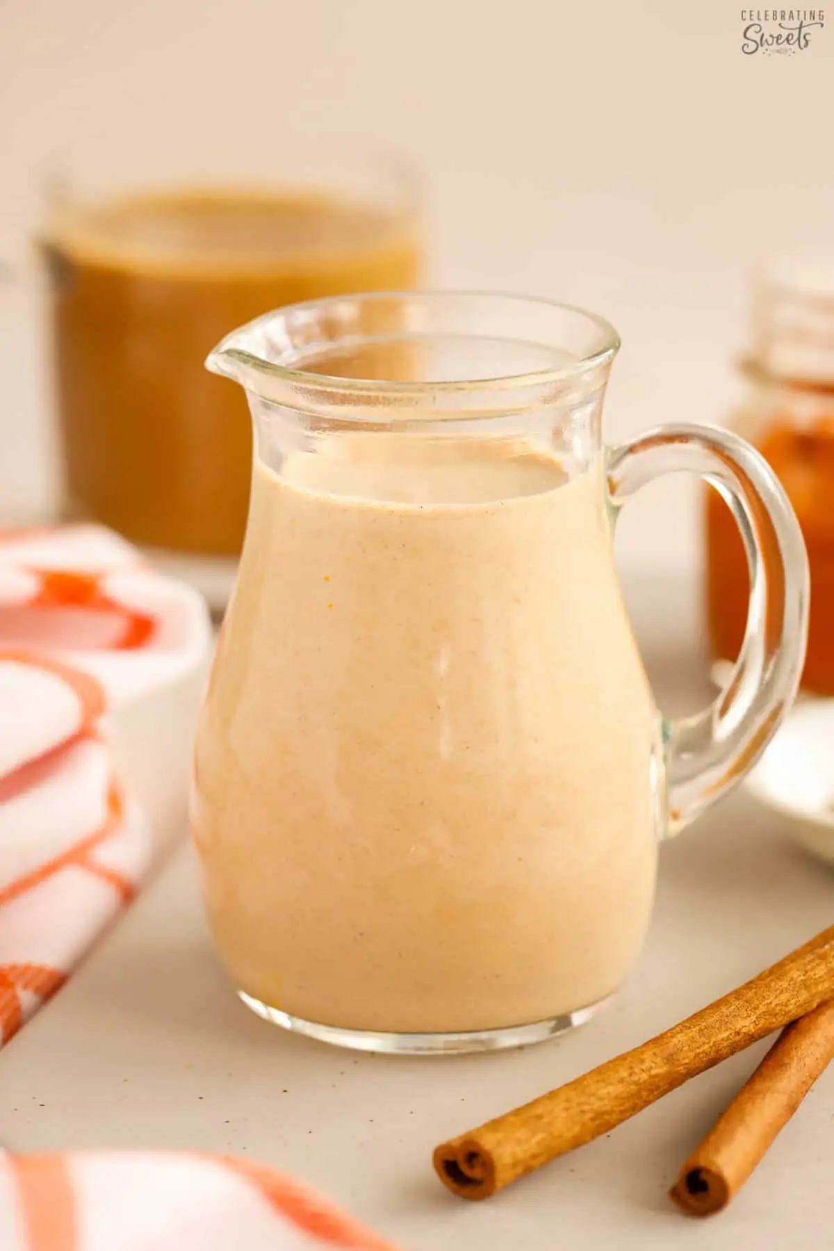 Pumpkin spice creamer in a glass carafe next to cinnamon sticks.