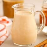 Pumpkin spice creamer in a glass carafe next to cinnamon sticks.