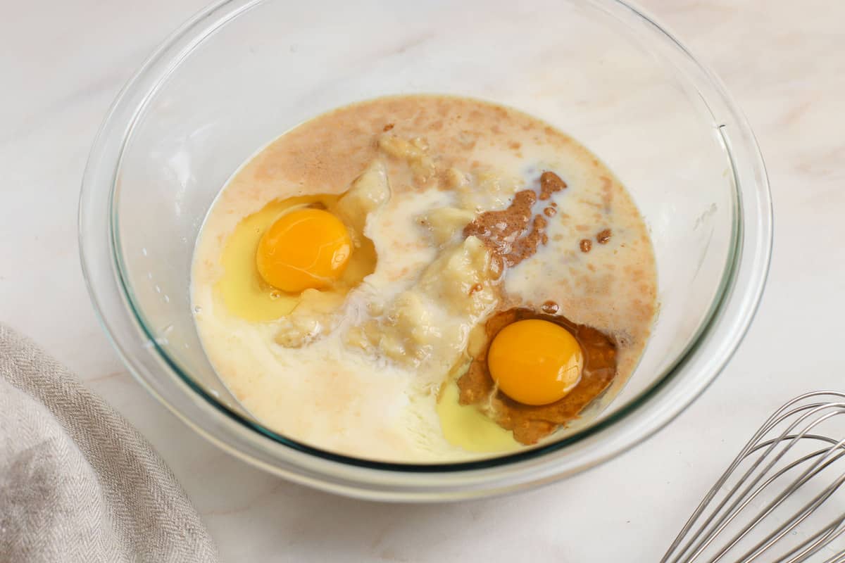 Eggs, banana, almond butter, milk in a glass bowl next to a whisk.
