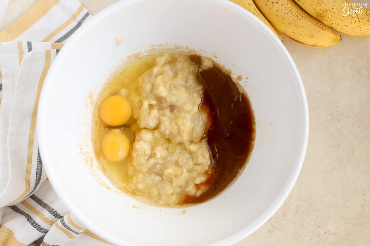 Mashed banana, brown sugar, eggs, and vanilla in a white bowl.