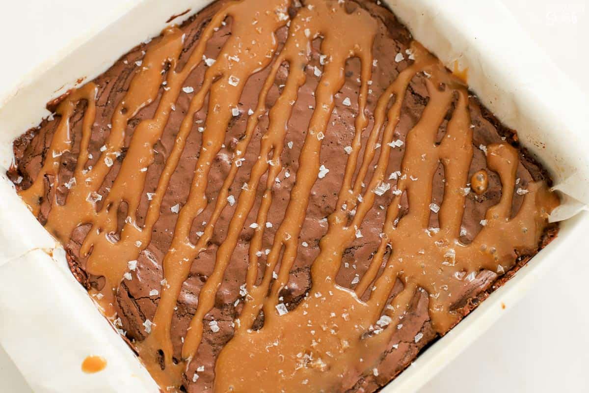 Brownies in a parchment-lined baking pan topped with drizzles of caramel sauce and flaky sea salt.