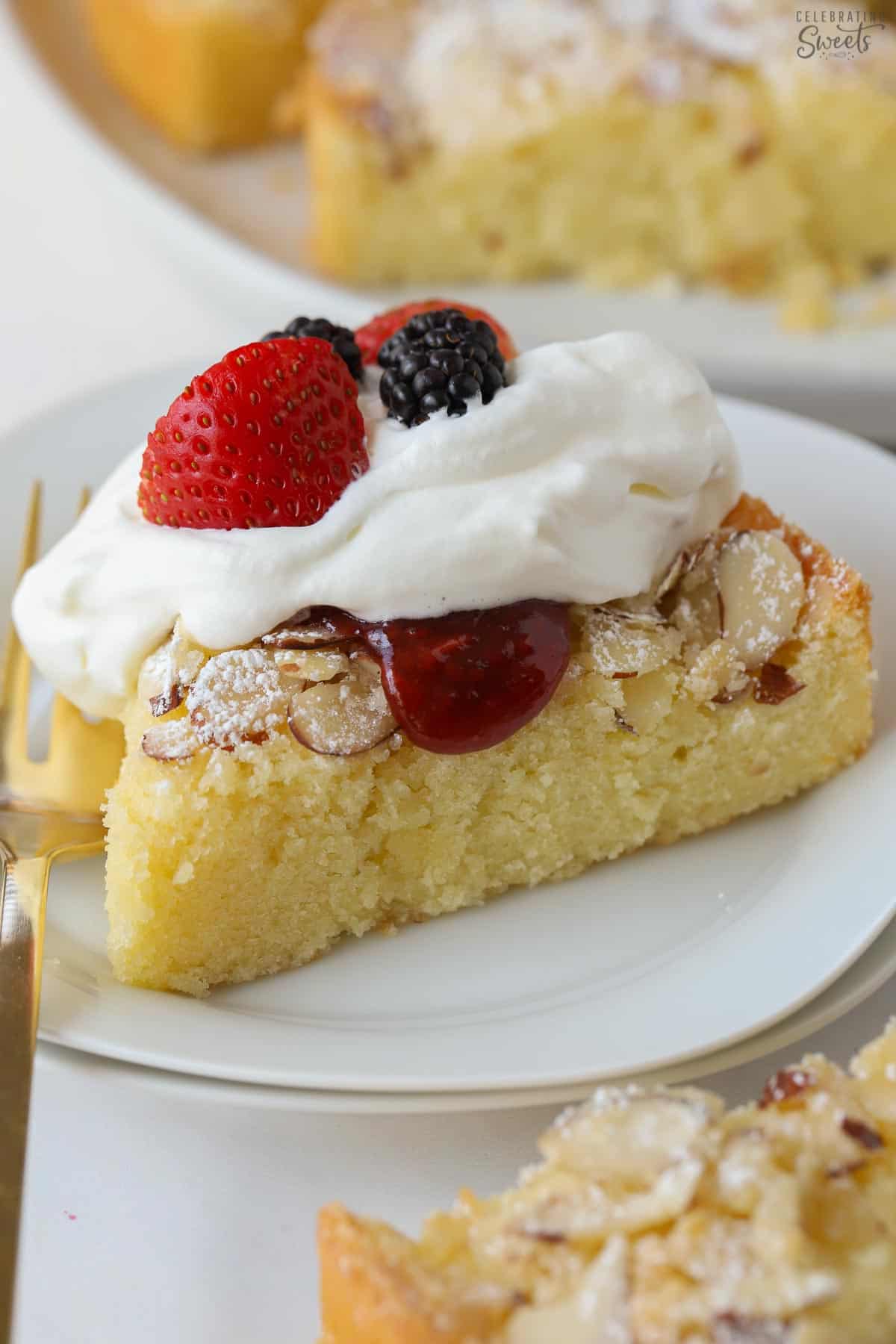 Almond cake with whipped cream and berries on a white plate with a gold fork.