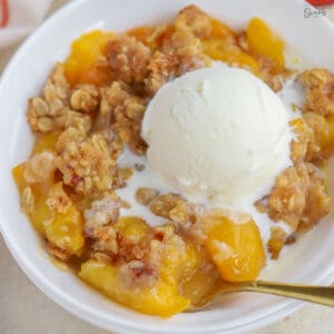 Peach crisp topped with vanilla ice cream in a white bowl.