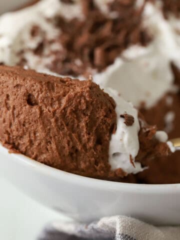 Gold spoon with chocolate mousse and whipped cream sitting on the edge of a white bowl.