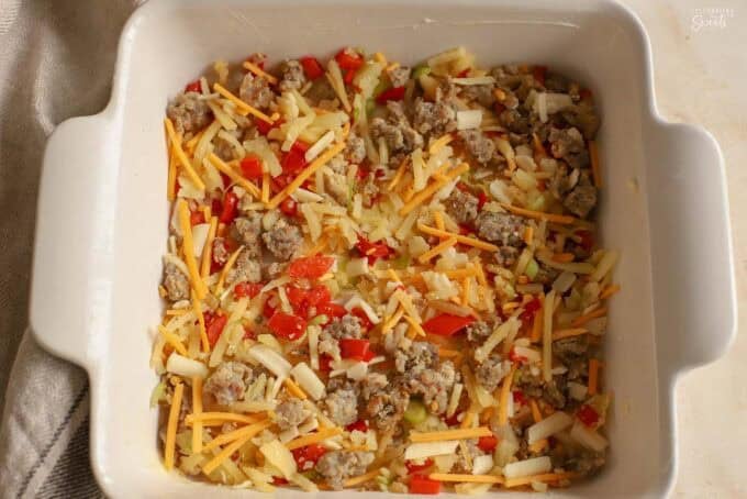 Hashbrowns, sausage, peppers, and cheese in a white square baking dish.