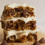 Stack of three oatmeal chocolate chip bars topped with white frosting surrounded by four large chocolate chips.