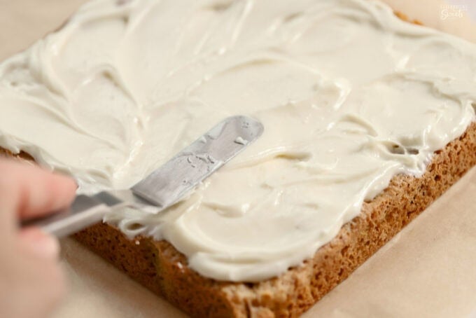 White frosting being spread on cookie bars sitting on brown parchment paper.