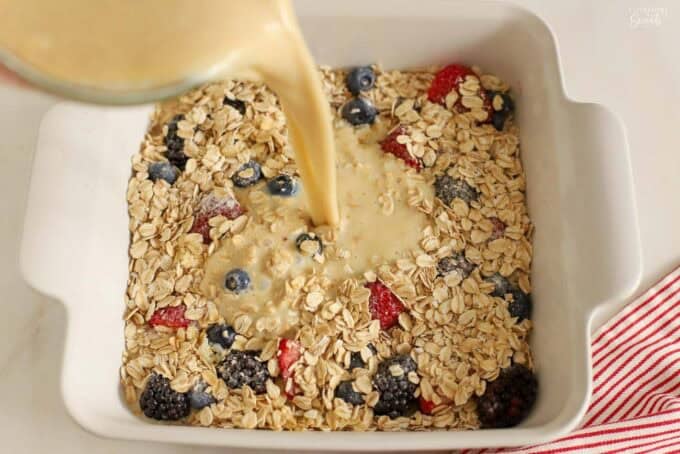 Liquid being poured into a white baking dish filled with berries and oats.