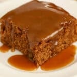 Slice of gingerbread cake on a white plate topped with toffee sauce.