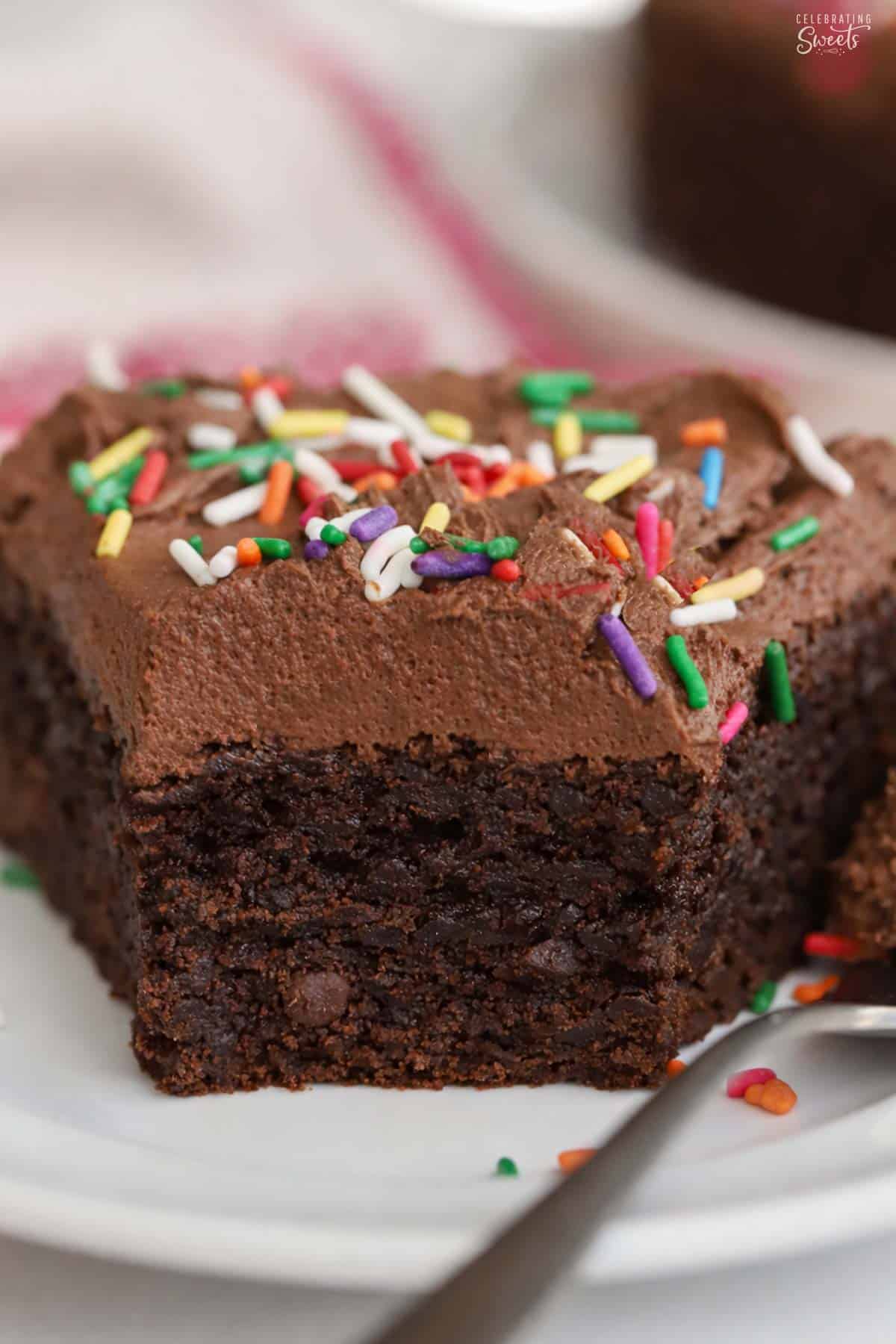 A slice of brownie cake topped with chocolate frosting and rainbow sprinkles on a white plate.