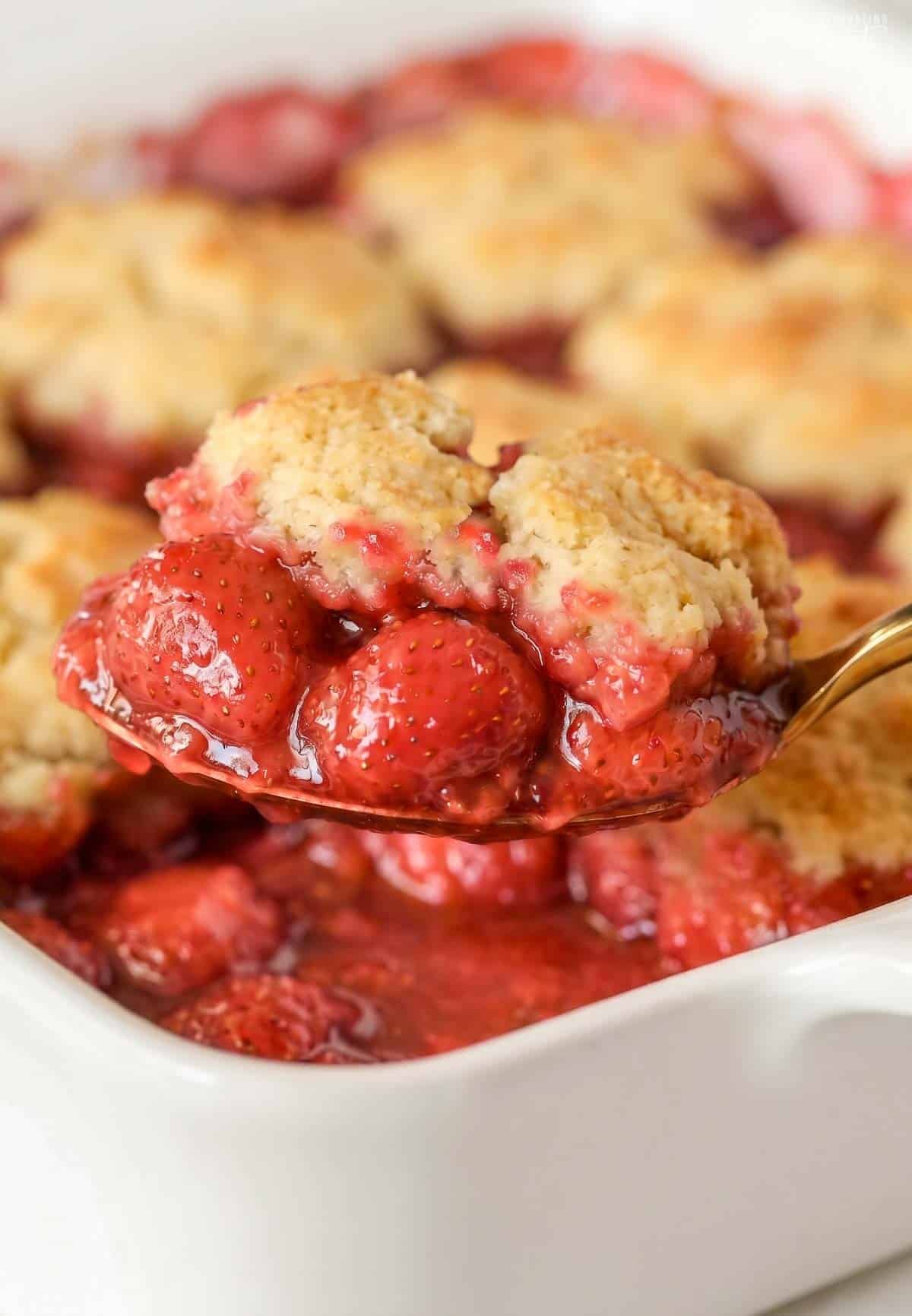 A scoop of strawberry cobbler on a gold spoon over a white baking dish.