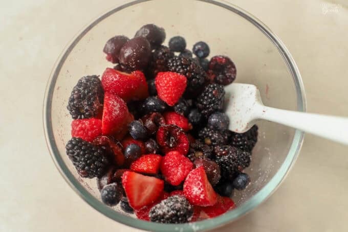 Fresh cherries and mixed berries in a glass bowl with a grey spatula.