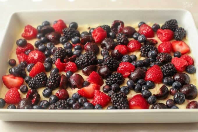 Fresh berries and cherries on top of vanilla cake batter in a white baking pan.