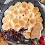 S'mores dip in a cast iron skillet next to graham crackers and strawberries.