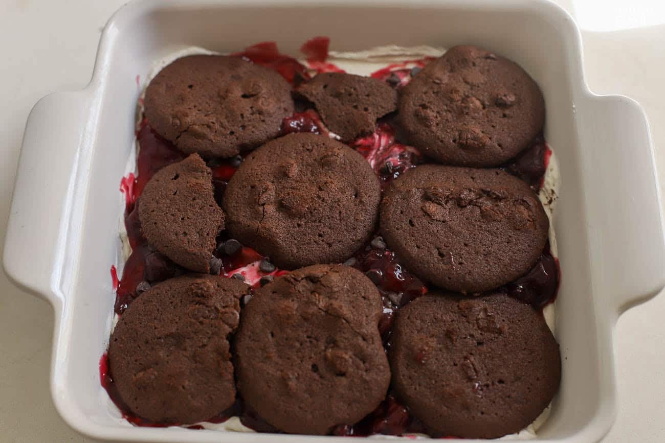 Black cherry icebox cake (chocolate cookies, whipped cream, and cherries) being assembled in a white square baking pan.